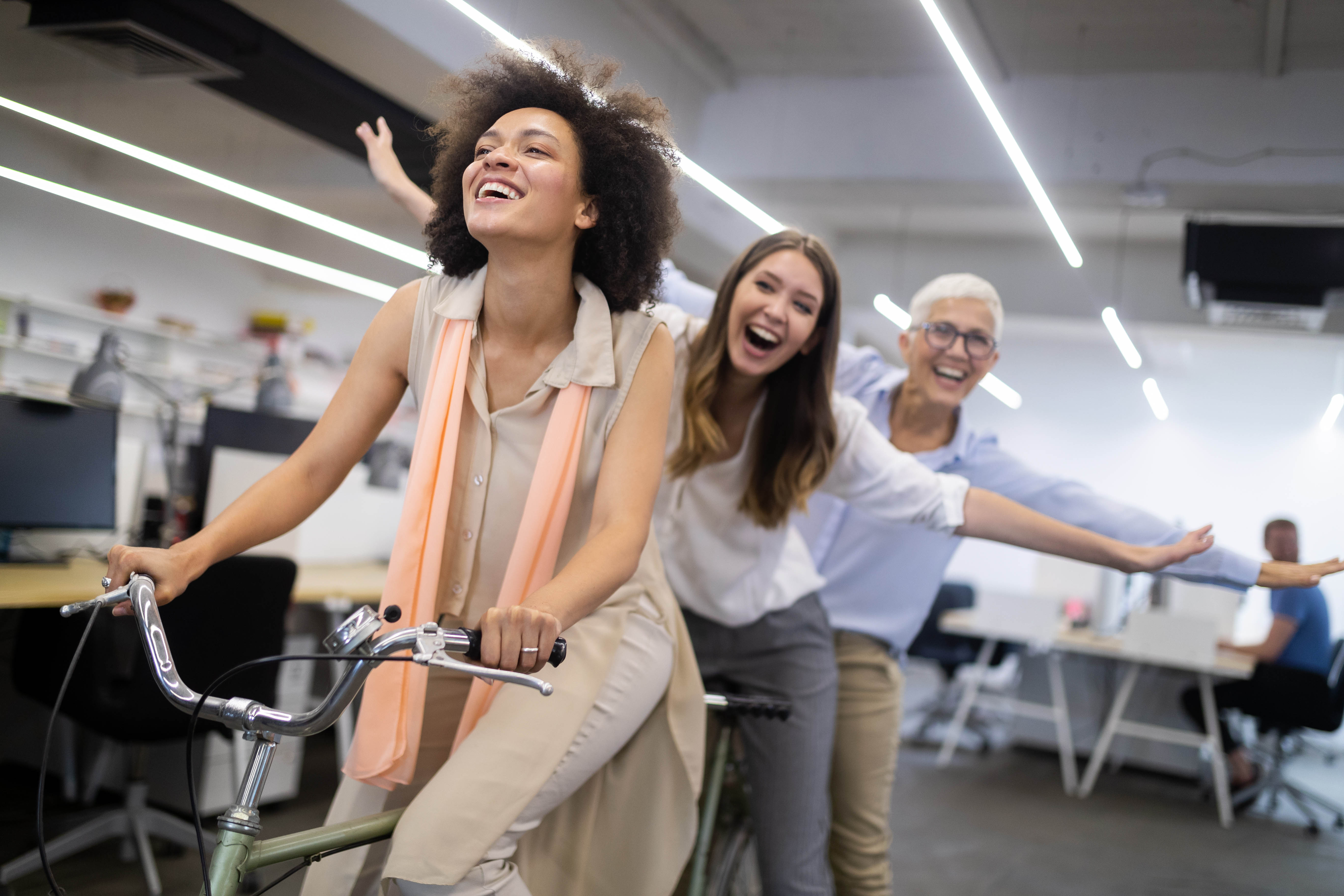 3 women of various ages riding a bike and laughing through the office, the two women on the back of the bike have their arms wide like they are flying